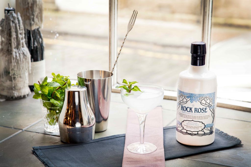 Bottle of Rock Rose Gin and Southwinds cocktail served in a coupe glass, with cocktail shaker and min sprigs