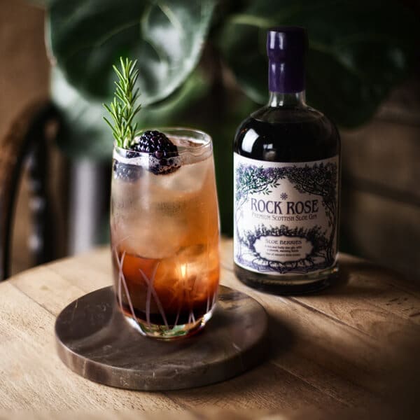 Bottle of Rock Rose Sloe Gin and perfect serve in a tall glass garnished with blackberries and sprig of thyme