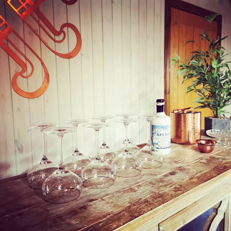 Branded coupe glasses and bottle of Rock Rose Gin displayed in on a wooden table in the tasting room