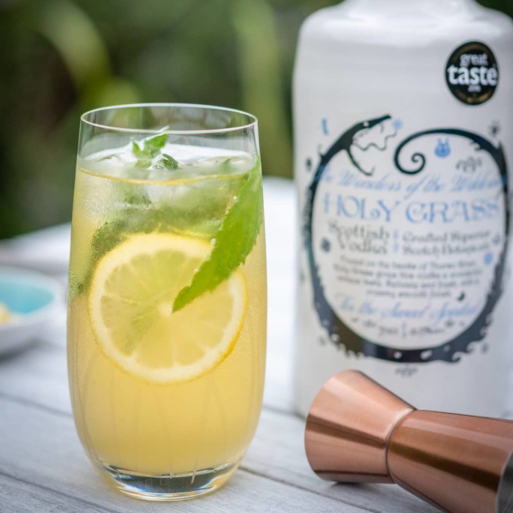 Bottle of Holy Grass Vodka and Minty Apple Cooler Cocktail served in a tall glass and garnished with lemon slice and mint leaf