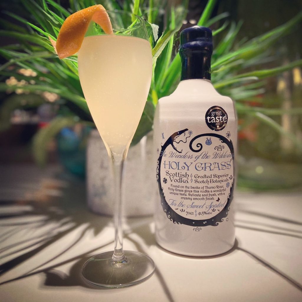 Bottle of Holy Grass Vodka and Peachy 5 Fizz cocktail served in a flute glass and garnished with orange peel