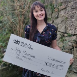 Ruth Potts posing outside in from of a stone wall holding a cheque of 3000 pounds for Help Musicians