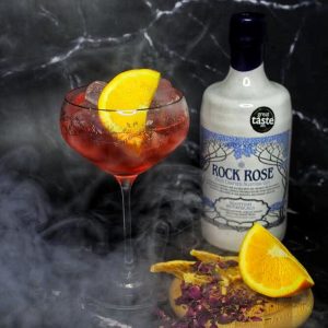 Bottle of Rock Rose Gin with Rose-Groni cocktail - by Mariusz Plucinski - served in a coupe glass and garnished with a slice of orange