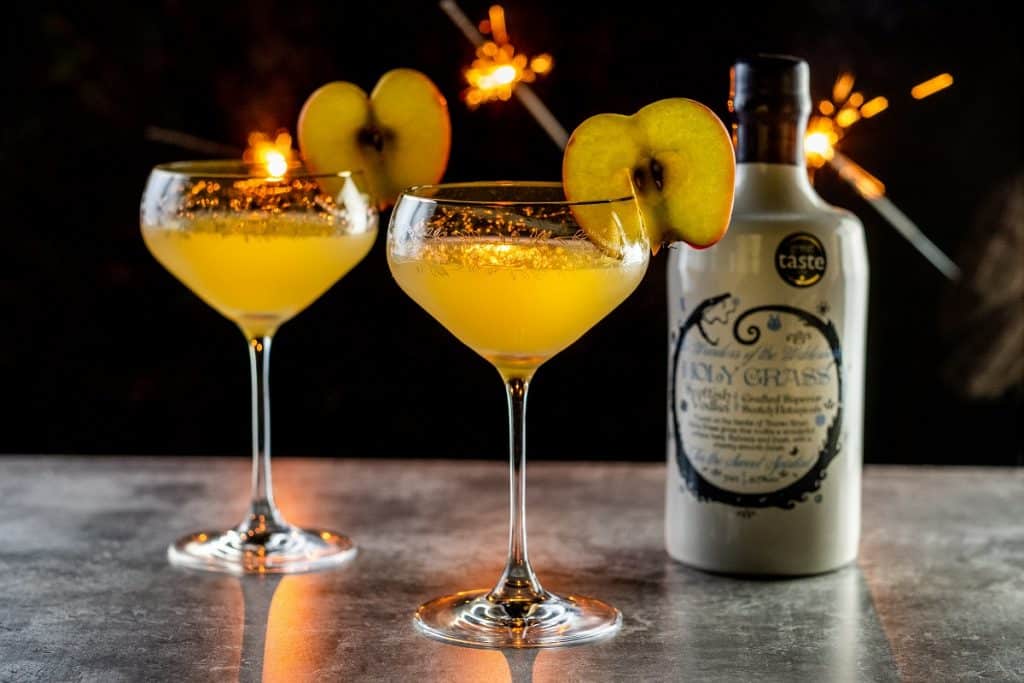 Bottle of Holy Gras Vodka and Toffee Apple-tini cocktail served in two coupe glasses and garnished with a slice of apple