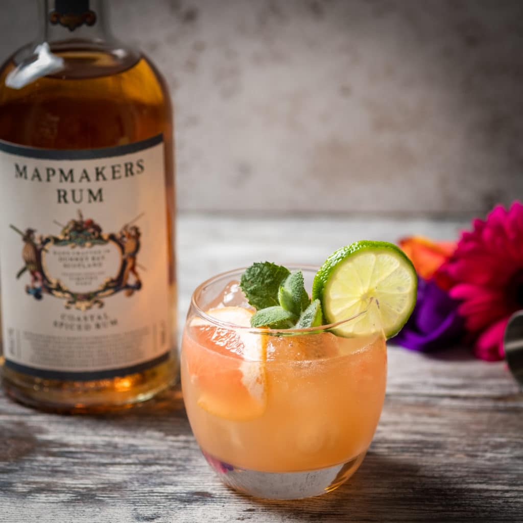 Bottle of Coastal Spiced Mapmaker's Rum with small glass of Mapmaker's punch garnished with a slice of pink grapefruit, lime wheel and mint sprig