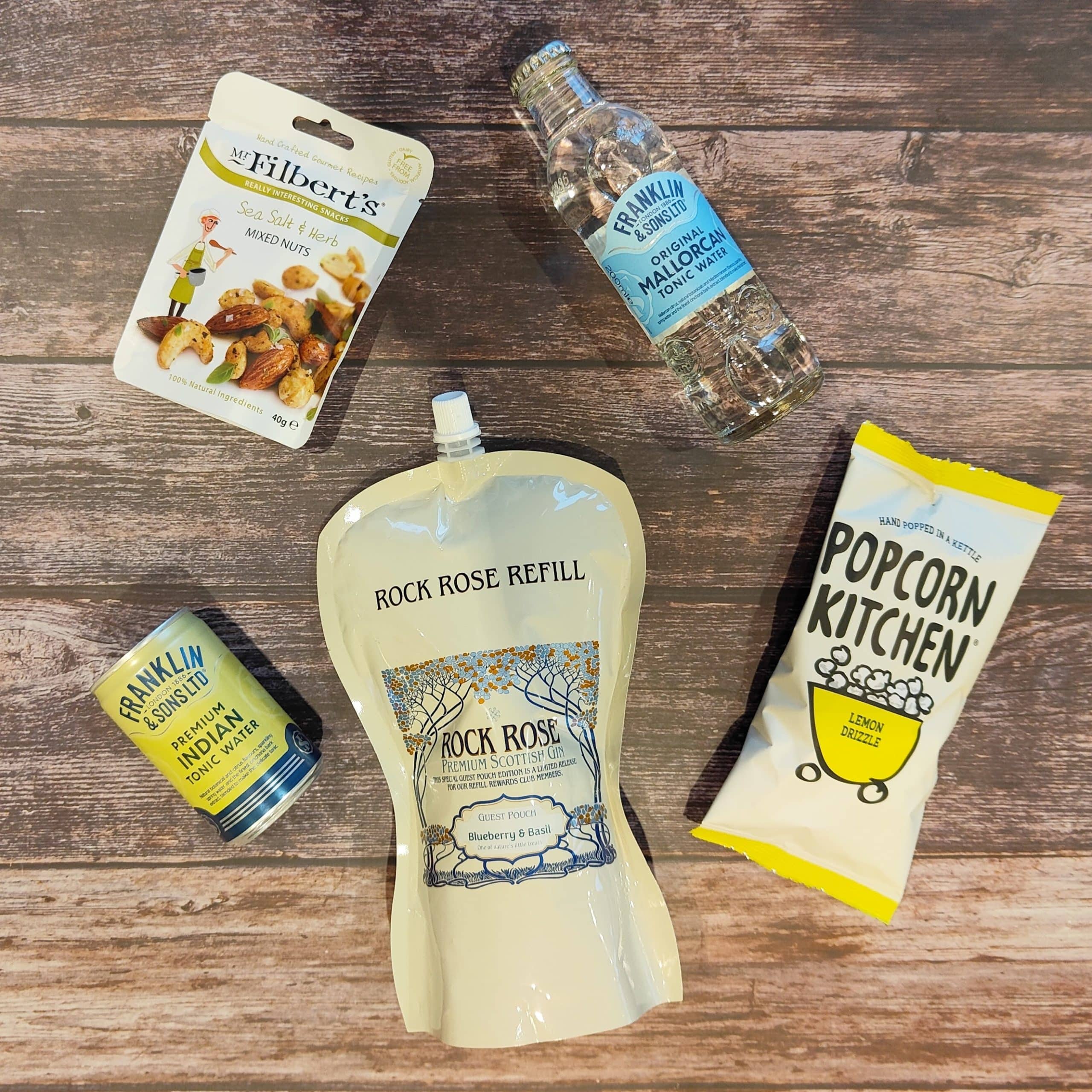 Content of the Refill Reward Club box for August 2023 including Rock Rose Gin Blueberry & Basil Guest pouch, 2 Tonic waters, Mixed nuts, Lemon Drizzle Popcorn